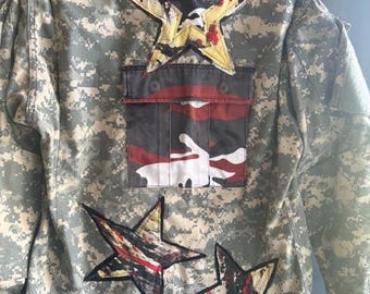 May include: A camouflage jacket featuring a digital pixelated pattern in shades of green, tan, and grey. The back is decorated with three star-shaped patches with black, yellow, and red paint splatters and a central pocket with red camo.