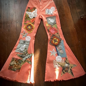 May include: Coral-colored flare jeans featuring a variety of decorative patches including crochet squares, white lace butterflies, star-shaped fabric appliqués, floral patterns, and small pockets. The hem of the pants is frayed and raw-edged.