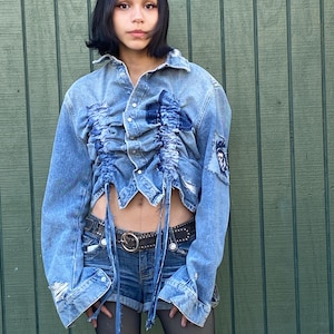 Puede incluir: Chaqueta vaquera azul claro con detalles desgastados y fruncidos. La chaqueta se combina con pantalones cortos vaqueros y un cinturón negro. El conjunto se completa con medias negras. Hay un parche en la manga.