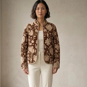 Può includere: Giacca trapuntata marrone con motivo floreale color crema e beige. La giacca ha un collo alto, due tasche anteriori e finiture beige. La modella indossa una canotta bianca e pantaloni color crema.