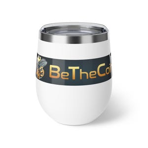 Puede incluir: Vaso de vino sin tallo blanco con borde y tapa plateados. El vaso presenta una banda negra con el texto dorado "BeTheCoin" y un gráfico de abeja.