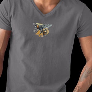 May include: A gray V-neck t-shirt featuring a detailed illustration of a bee with yellow and black stripes. The bee is positioned above a yellow number 3. The shirt is made of a soft material.