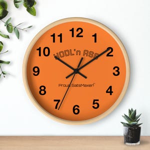 May include: Round wall clock with an orange face and black numerals. The clock has a light wood frame and black hands. The text "HODL'n ASS" and "Proud SatsMaxer!" are printed on the clock face.