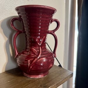 May include: A maroon ceramic vase with two handles on each side. The vase has a ribbed texture and a decorative leaf design. It sits on a wooden shelf. The vase is a decorative item.