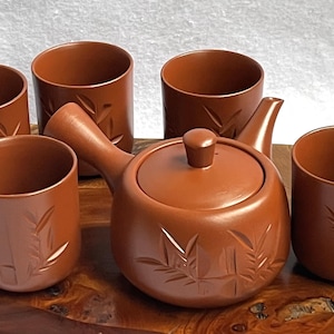 Peut inclure: Un service à thé en céramique marron comprenant une théière et quatre tasses, chacune ornée d'un motif de feuilles de bambou. La théière a un couvercle et une anse. L'ensemble est présenté sur un plateau en bois.