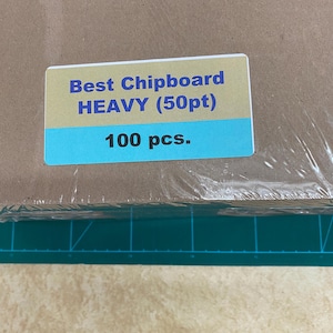 May include: A stack of brown chipboard sheets, wrapped in clear plastic. A rectangular label with the text "Best Chipboard HEAVY (50pt) 100 pcs." is affixed to the packaging. The label has a gold top and a blue bottom.