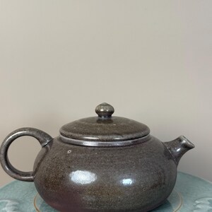 May include: A brown ceramic teapot with a lid and a curved handle. The teapot has a spout and a small knob on top of the lid. The teapot is sitting on a light blue surface.