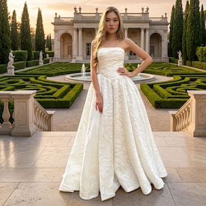 Könnte beinhalten: Ein trägerloses elfenbeinfarbenes Hochzeitskleid mit weitem Rock und dezentem floralem Muster. Das Kleid hat ein eng anliegendes Oberteil und einen fließenden Rock. Im Hintergrund ein formeller Garten, ein Brunnen und ein großes Gebäude.