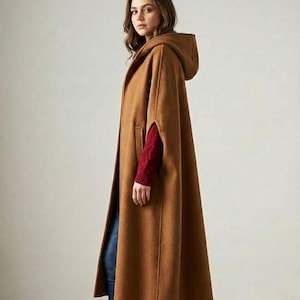 May include: A long, hooded cape in a warm brown color. The cape features a flowing design with arm openings and a hood. The model is wearing a red sweater underneath. The cape is made of a soft material.