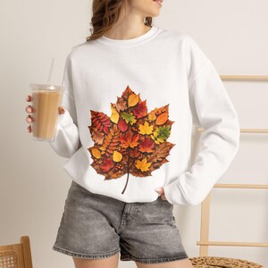 May include: A white long-sleeve sweatshirt featuring a large, central graphic of a single maple leaf composed of a collage of smaller autumn leaves in shades of red, orange, yellow, brown, and green. The sweatshirt has a crew neckline and ribbed cuffs.