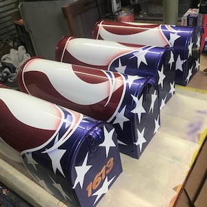 May include: Four mailboxes painted with an American flag design. The mailboxes are blue with white stars and red and white stripes. The mailbox on the right has the number 1673 painted on it.