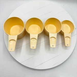 Può includere: Quattro misurini giallo chiaro con manici sono disposti su una superficie bianca. Le tazze sono in plastica e hanno una finitura liscia e opaca. I manici hanno un foro all'estremità per appenderli. Le tazze sono nidificate.