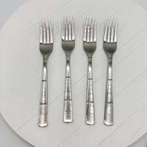 Può includere: Quattro forchette d'argento dal design semplice ed elegante. Ogni forchetta ha un manico lungo e affusolato con una fascia decorativa vicino alla parte superiore. I rebbi sono distanziati uniformemente e leggermente curvi. Le forchette sono disposte su una superficie bianca.