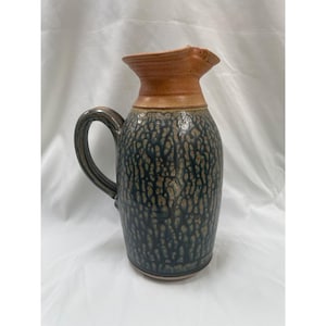 May include: A ceramic pitcher with a dark blue and tan speckled pattern. The pitcher has a brown rim and handle. The handle is a dark brown color. The pitcher is a decorative item.