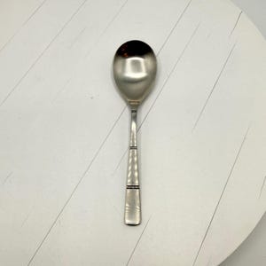 Può includere: Un cucchiaio d'argento con una ciotola arrotondata e un lungo manico rettangolare. Il manico presenta un design decorativo con due sottili bande nere. Il cucchiaio è realizzato in metallo lucido e riflettente.