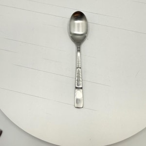 Può includere: Un cucchiaio d'argento con una coppa arrotondata e un manico strutturato. Il manico ha una forma rettangolare con due fasce decorative. Il cucchiaio è posto su uno sfondo bianco.