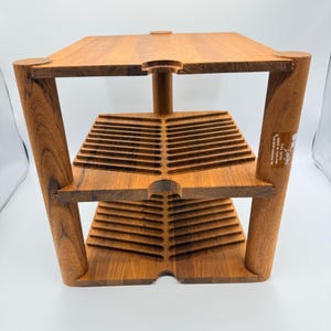 Może przedstawiać: Trzypoziomowa drewniana półka z naturalnym wykończeniem drewna. Półka ma prostokątny blat i dwie dolne półki z rowkowanymi detalami. Podpory są zaokrąglone i ustawione pod kątem. Półka ma około 30 cm szerokości, 30 cm głębokości i 30 cm wysokości.