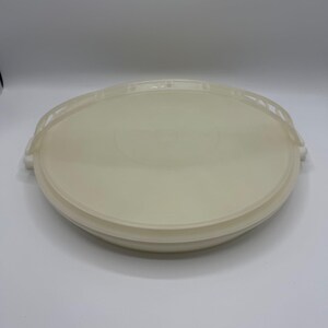 May include: A round, off-white plastic container with a matching lid. The lid has a handle on each side and a slightly raised center. The container appears to be designed for food storage.