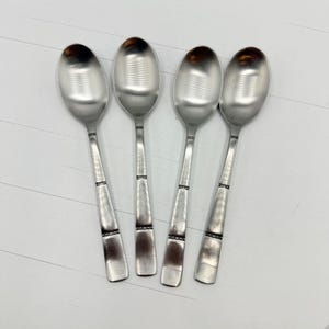 Può includere: Quattro cucchiai d'argento con coppette ovali e manici lunghi e affusolati. I manici hanno una fascia decorativa vicino alla base della coppetta e un'altra vicino all'estremità. I cucchiai sono disposti su una superficie bianca.