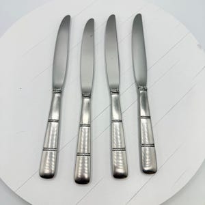 Può includere: Quattro coltelli da burro in acciaio inossidabile disposti su una superficie bianca e rotonda. I coltelli hanno un manico strutturato e una fascia decorativa. Le lame sono lisce e lucenti. Ideali per spalmare.