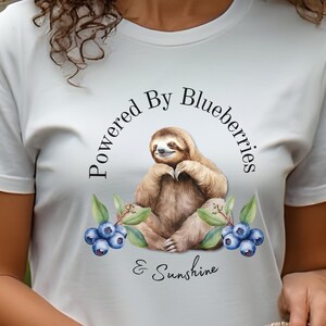 May include: A white t-shirt featuring a central graphic of a brown sloth sitting among green leaves and blue blueberries. The text "Powered By Blueberries & Sunshine" is printed in black lettering around the sloth illustration on the front.