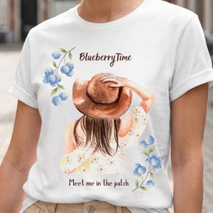 May include: White t-shirt with a watercolor illustration of a woman wearing a brown hat, with the text "Blueberry Time" and "Meet me in the patch". The design includes blue watercolor blueberries and flowers.