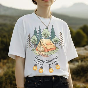 May include: A white short-sleeve t-shirt featuring a graphic illustration of an orange tent nestled among green trees. Below the tent, the text "Happy Camper" is written in black, accompanied by a string of four glowing yellow light bulbs.