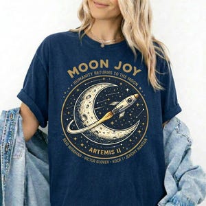 May include: Dark blue t-shirt with a circular graphic. The graphic shows a crescent moon, a rocket, and the text "MOON JOY" and "HUMANITY RETURNS TO THE MOON". The design also includes the text "ARTEMIS II" and the crew members' names.
