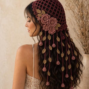 May include: A deep burgundy crochet head covering featuring a lattice pattern, adorned with three dimensional dusty pink floral appliqués and dangling strands of crocheted tan leaves and small pink flower buds that drape down the back.