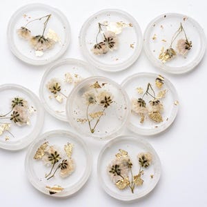May include: A collection of eight circular clear resin coasters, each featuring embedded dried white flowers and delicate gold leaf flakes. The coasters have a smooth, transparent finish with visible botanical details and metallic accents throughout.