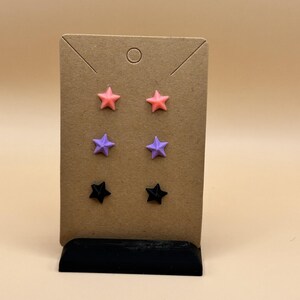 May include: A set of three pairs of star-shaped stud earrings in pink, purple, and black. The earrings are displayed on a brown card with a black base, set against a neutral background. The earrings are small and likely made of plastic or resin.