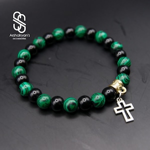 May include: A beaded bracelet featuring alternating black and green malachite stones. A gold-toned bead and a silver cross charm dangle from the bracelet. The brand name "Arshakyan's accessories" is in the upper left corner.