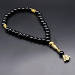 Peut inclure: Chapelet de prière à perles noires avec des accents décoratifs dorés. Les perles sont enfilées ensemble, formant une boucle. Les accents dorés comprennent des pièces d'extrémité et des séparateurs ornés. Les perles sont probablement en pierre polie.