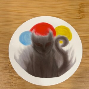 May include: A round sticker featuring a stylized, shadowy figure with a red head covering and a curled tail. The design includes blue and yellow orbs, creating a whimsical and artistic aesthetic. The sticker is on a wooden surface.