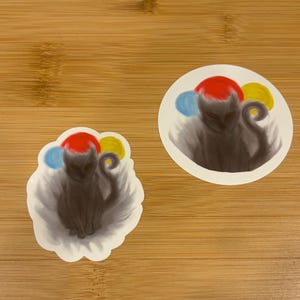 May include: Two stickers featuring a dark gray creature with a red head, blue and yellow accents, and a curled tail. One sticker is round, the other is cloud-shaped, both with a white border. The stickers are on a wooden surface.