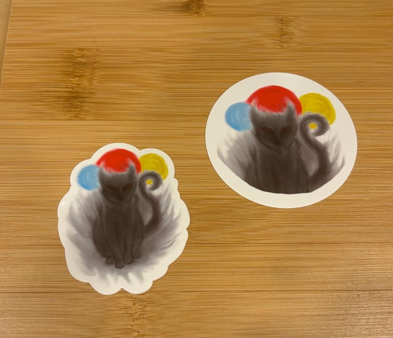May include: Two stickers featuring a dark gray creature with a red head, blue and yellow accents, and a curled tail. One sticker is round, the other is cloud-shaped, both with a white border. The stickers are on a wooden surface.