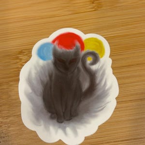 May include: A sticker featuring a stylized black cat with a red hat and three colorful orbs. The cat is sitting, with its tail curled. The sticker has a white, cloud-like border and is on a wooden surface.