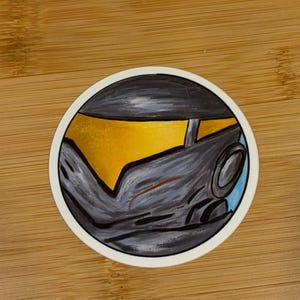 May include: Round sticker featuring a stylized illustration of a helmet. The design includes shades of gray, yellow, and blue, with a red accent. The sticker has a white border and is likely a decorative item.