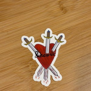 May include: A sticker depicting a red heart pierced by three swords. The heart has blood dripping from it, and the words "& hearts" are written across it in a handwritten font. The swords have silver blades and gold hilts.