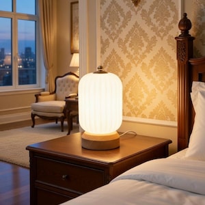 May include: A white, ribbed glass table lamp with a wooden base sits on a dark brown nightstand. The lamp is illuminated, casting a warm glow in a bedroom setting. The room features a window with a city view.