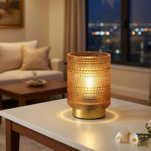 May include: A cylindrical amber glass lamp with a textured surface and a gold base. The lamp is lit, casting a warm glow on the white table it sits on. The background includes a sofa, coffee table, and a window with city lights.