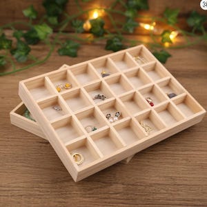 Pode incluir: Duas bandejas de joias de madeira clara com múltiplos compartimentos. As bandejas estão cheias de várias peças de joias, incluindo anéis, brincos e colares. As bandejas estão empilhadas uma sobre a outra, com a bandeja superior ligeiramente inclinada.