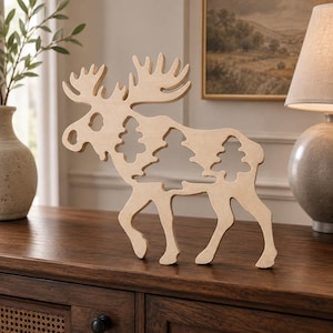 May include: A light-colored wooden cutout of a moose standing in profile. The body of the moose features intricate, hollowed-out silhouettes of three evergreen trees, creating a decorative cutout design with smooth, natural wood grain edges.