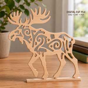 May include: A light-colored wooden cutout of a moose featuring intricate scrollwork patterns throughout its body and antlers. The design stands on a rectangular base. Text in the upper right corner reads: DIGITAL CUT FILE SVG DXF PDF.