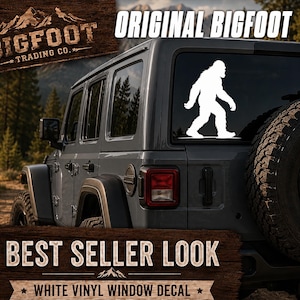 Op de afbeelding: Een witte vinyl raamsticker met een silhouet van een lopende Bigfoot, aangebracht op de achterruit van een grijze SUV. De sticker is een effen witte afbeelding die het legendarische wezen in profiel op het voertuigvenster toont.