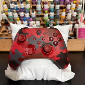 May include: A wireless video game controller featuring a red, dark red, and black camouflage pattern. The controller includes two analog sticks, a directional pad, four action buttons labeled A, B, X, and Y, and a central Xbox logo button.