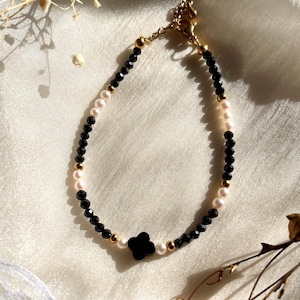 May include: A beaded bracelet featuring alternating black faceted beads and small white pearls, accented with gold-toned metal beads. A central black four-leaf clover charm is positioned at the bottom, and the bracelet includes a gold-toned metal clasp.
