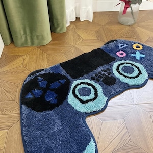Könnte beinhalten: Eine blaue, plüschige Badematte in Form eines Videospiel-Controllers. Die Matte hat schwarze, hellblaue und türkisfarbene Akzente mit kreisförmigen und Richtungstasten-Designs. Die Matte liegt auf einem Holzboden.