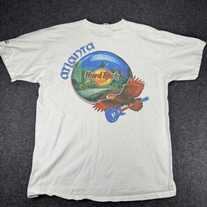 Könnte beinhalten: Weißes T-Shirt mit einem Aufdruck eines Adlers, der eine blaue Gitarre spielt, mit den Worten "Atlanta" und "Hard Rock Cafe" in einem kreisförmigen Design. Der Adler hat rote und braune Federn.