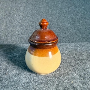 May include: A small, ceramic jar with a lid. The jar is a light beige color on the bottom, transitioning to a band of orange, and topped with a dark brown lid. The lid has a small, pointed finial. The jar is likely for storing spices or other small items.